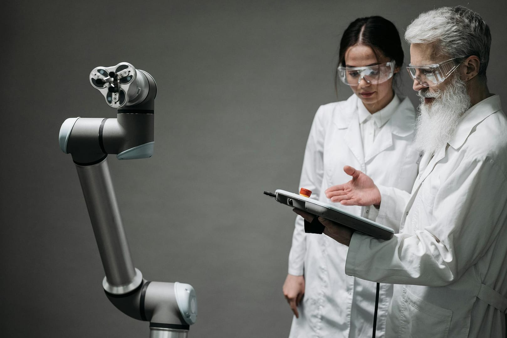 Researchers in lab coats and safety glasses engaging with a robotic arm in a lab setting.